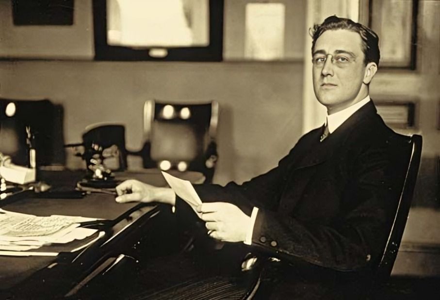 Young man in a suit seated at a desk, holding a paper, and looking thoughtfully to the side.