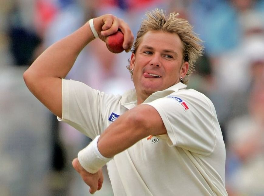 A cricketer in action, wearing white, prepares to bowl with a red ball in a lush, blur background.
