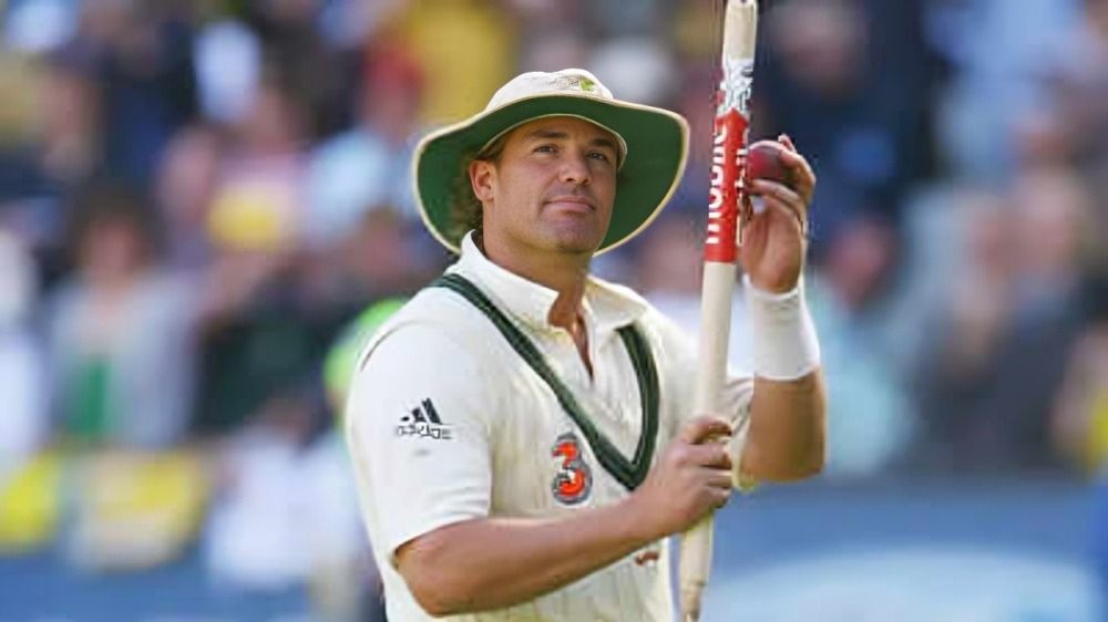 A cricketer in a white uniform and hat holds his bat up.