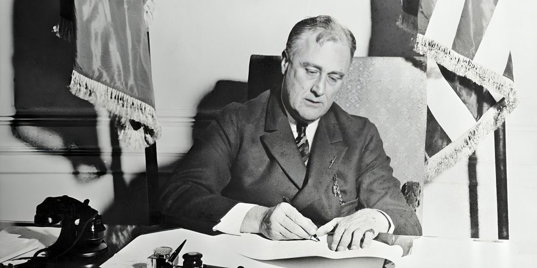 President Franklin D. Roosevelt signed a document at his desk, with flags in the background.