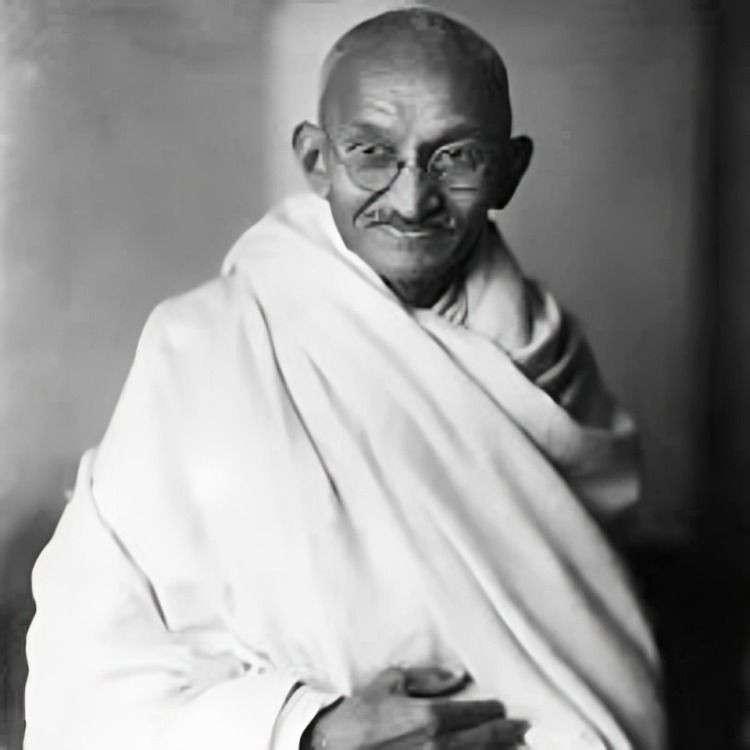 Portrait of Mahatma Gandhi with glasses, dressed in a simple white shawl against a dark background.