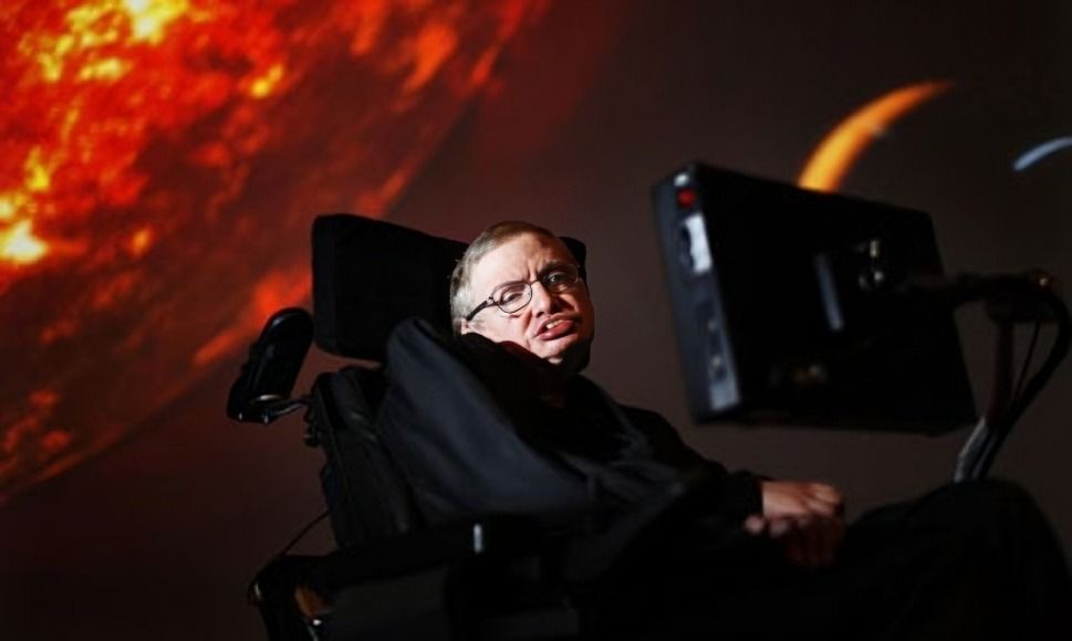 Stephen Hawking, seated in his wheelchair, in front of a screen, dark background.