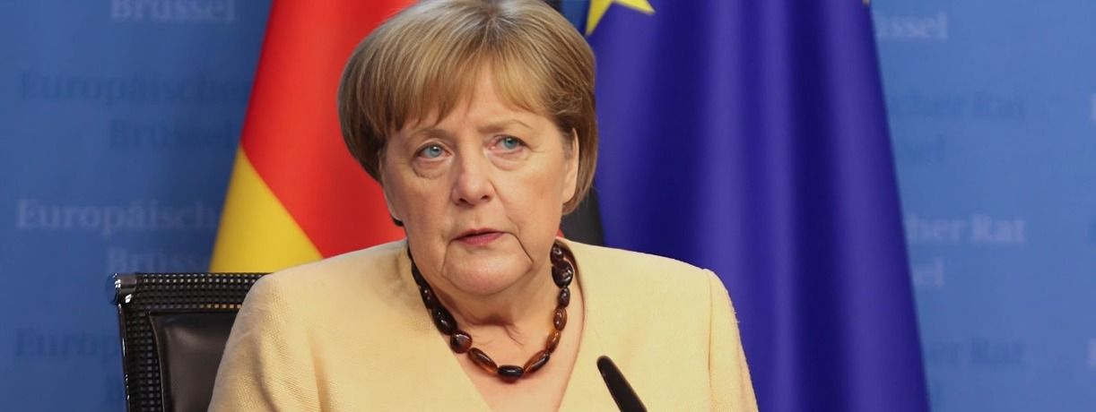 Angela Merkel speaking at a press conference, flags visible in the background.