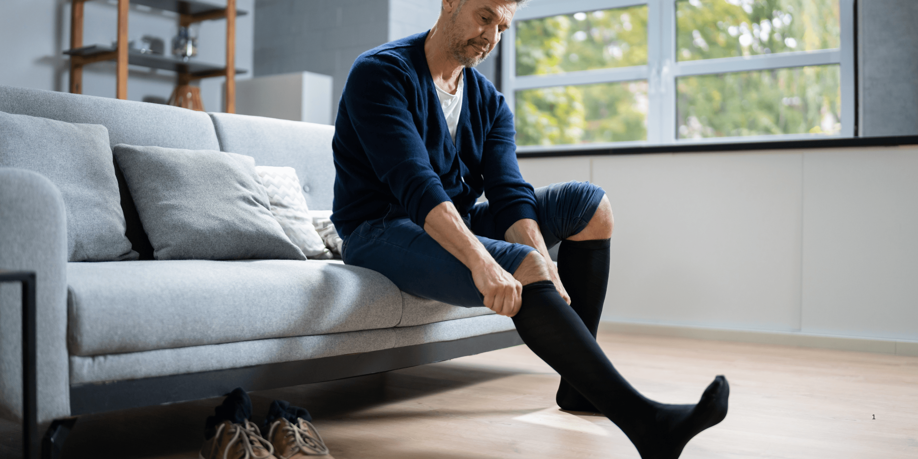 A man sitting on a couch putting on long black socks.