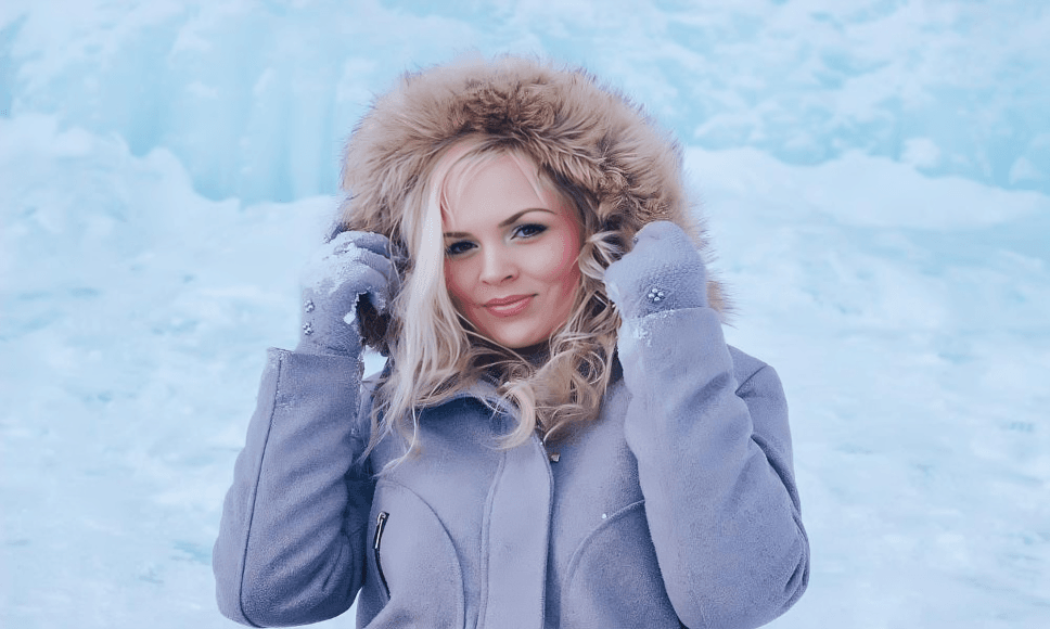 A blonde woman in a gray winter coat and mittens.