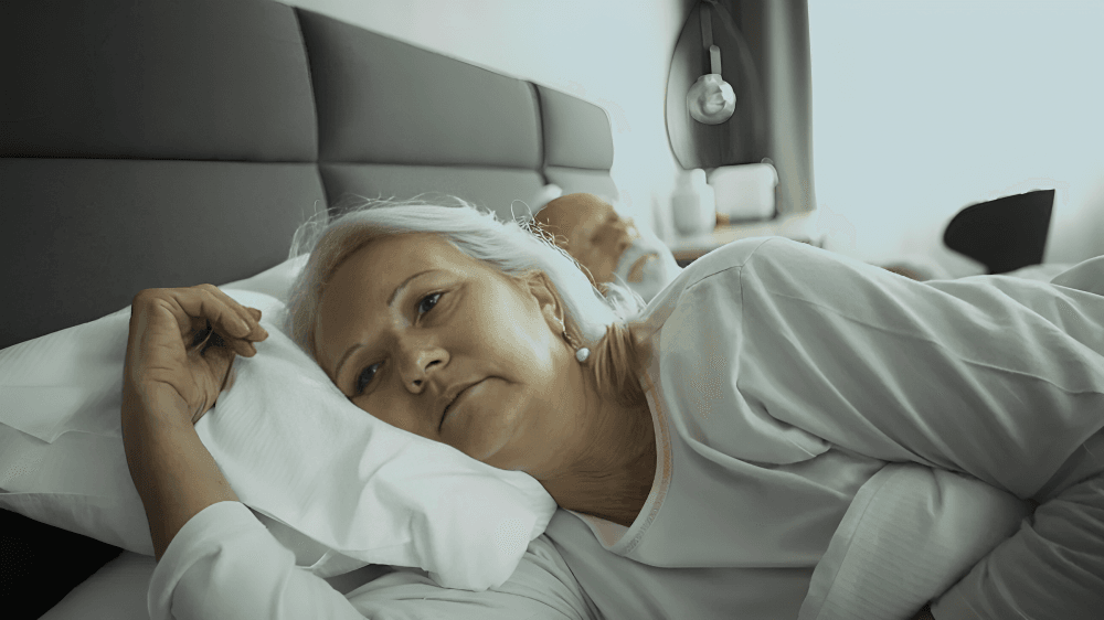 Senior woman looks thoughtful in bed, man behind her.