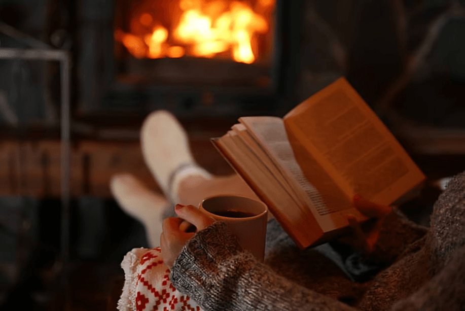 Person reading book by warm fireplace with drink.