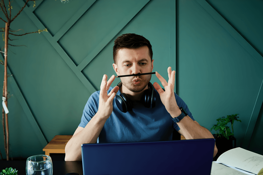 A man playfully holds a pencil-like mustache at a laptop.