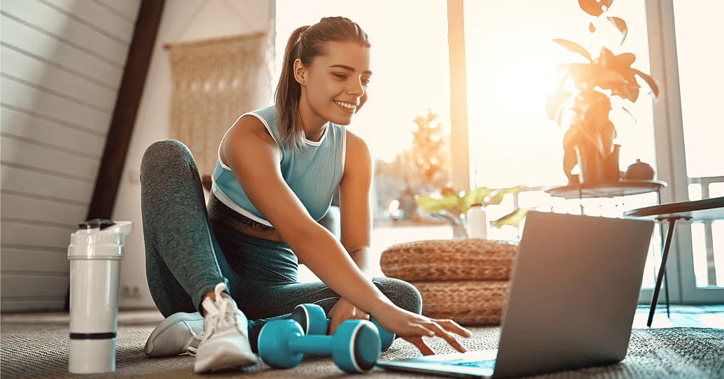 A smiling woman exercises at home with a laptop.