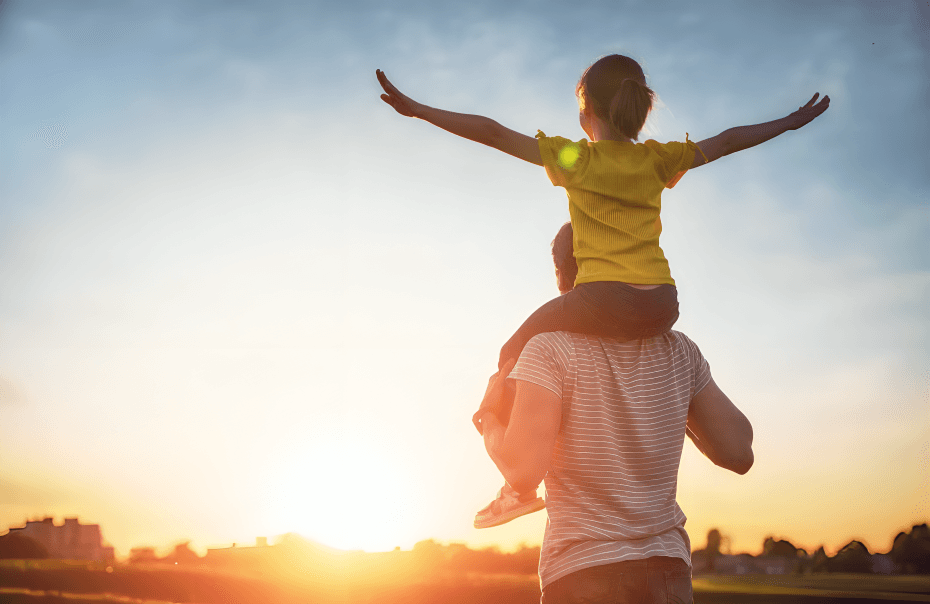 A child on shoulders at sunset, arms outstretched.