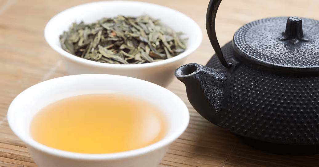 A cup of green tea with loose leaves and a teapot.