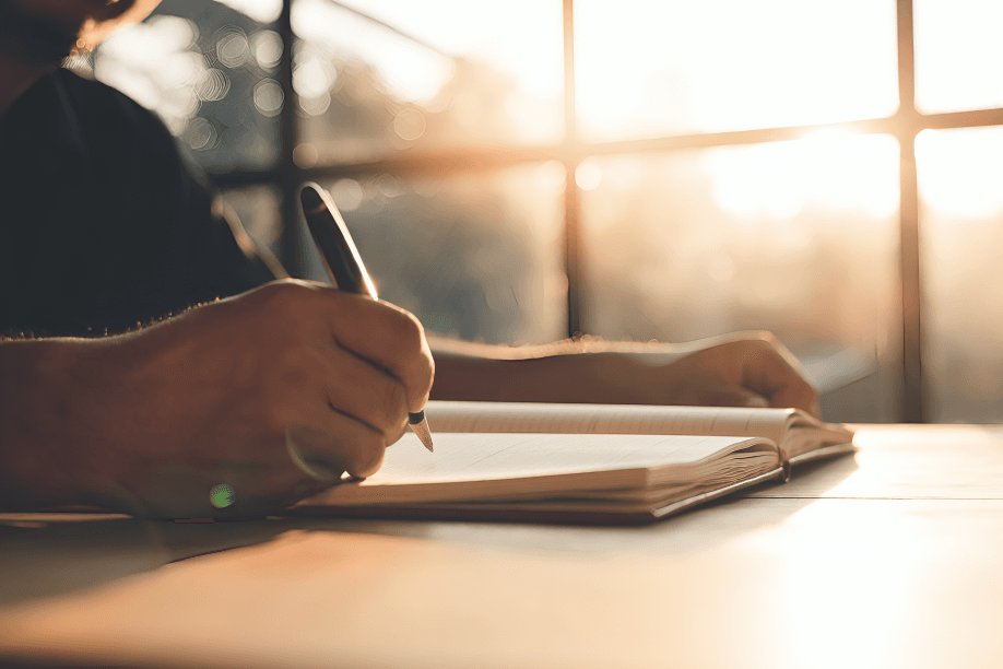 Hands writing in a journal by a bright window.