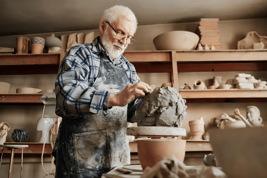 An elderly man sculpts clay in a pottery studio.