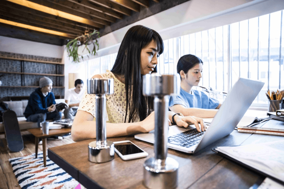 Two women, possibly of East Asian descent, work on laptops in a shared space.