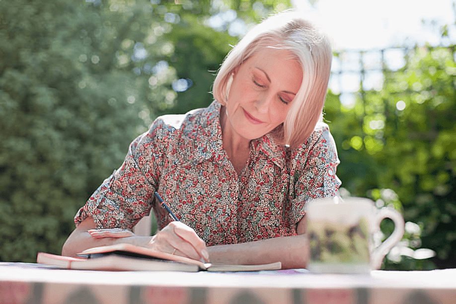 A woman writing in a notebook outdoors with a cup.