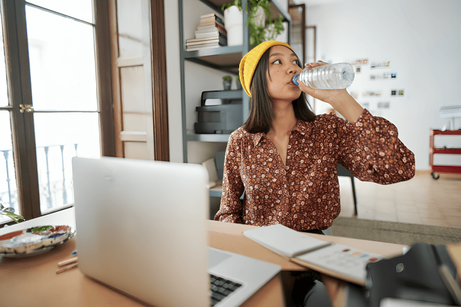 A young woman in a yellow beanie drinks water at a desk.