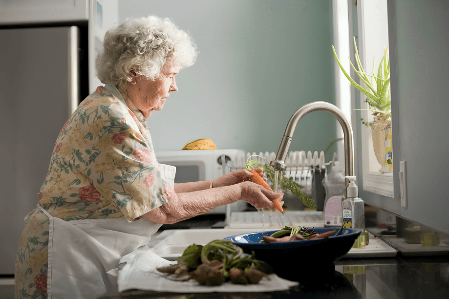 Elderly woman washes carrots in kitchen sink.