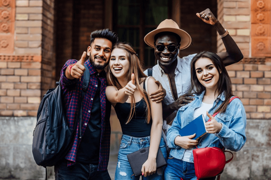 Four smiling young adults of diverse appearances give thumbs up.