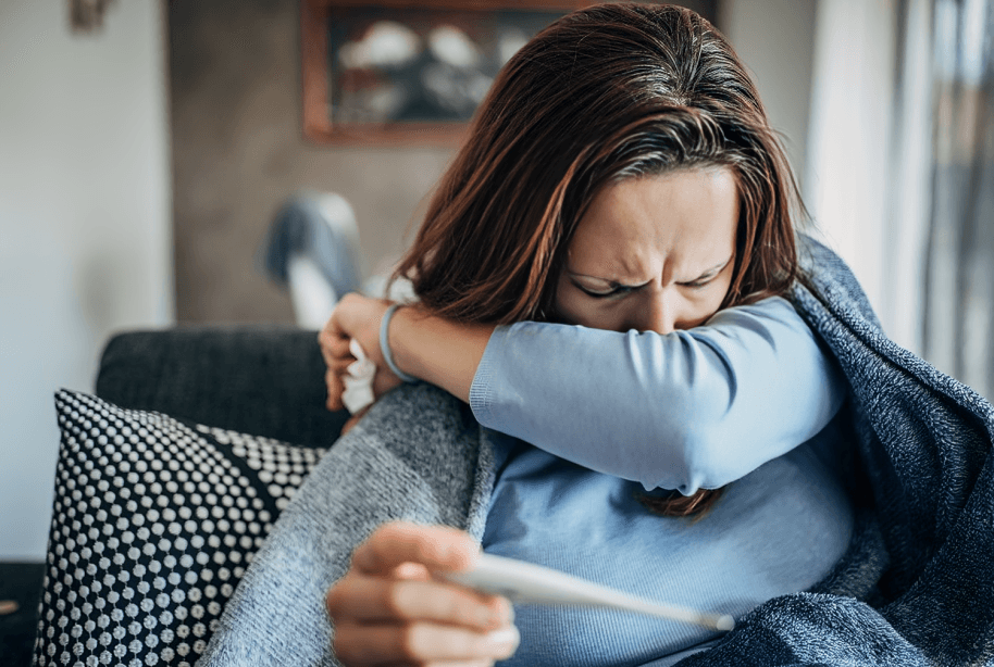 Woman feeling sick, coughing into elbow and holding thermometer.