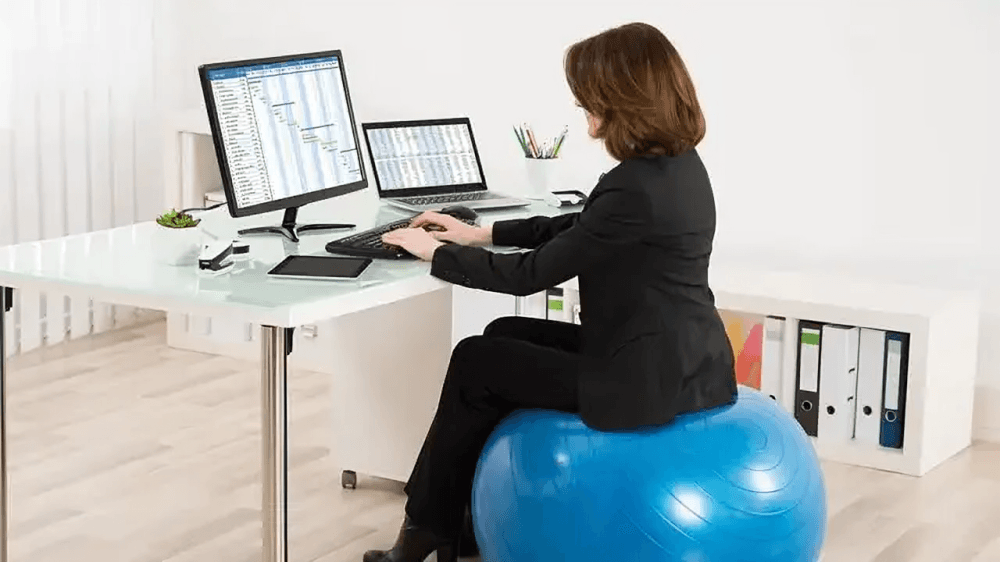 A woman works at desk, sitting on a blue exercise ball.