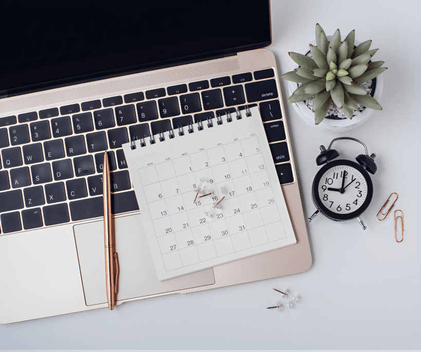Calendar, laptop, alarm clock, and plant on a desk.