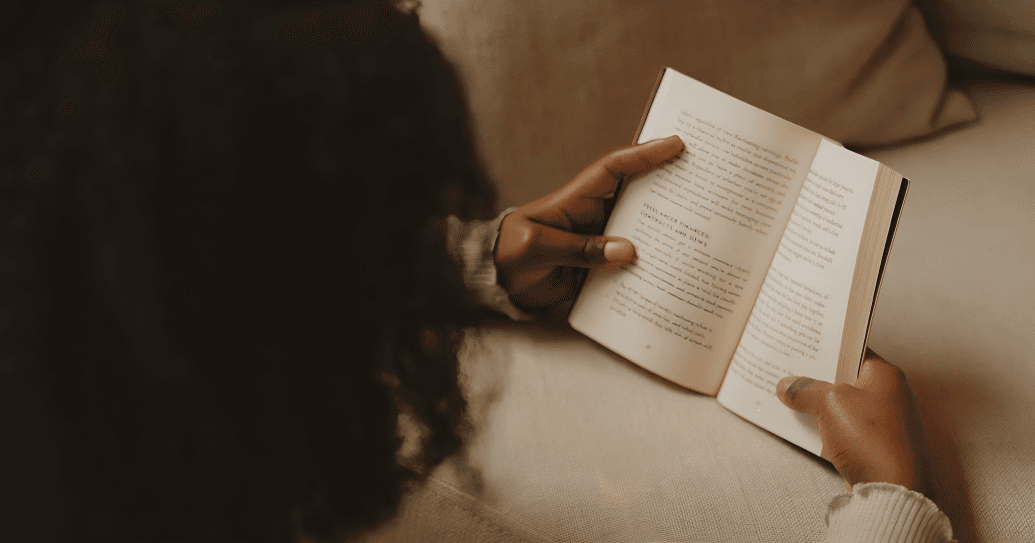 Person on couch, hands holding open book, reading.
