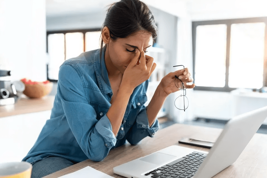 Stressed woman rubs eyes at laptop on bright desk.