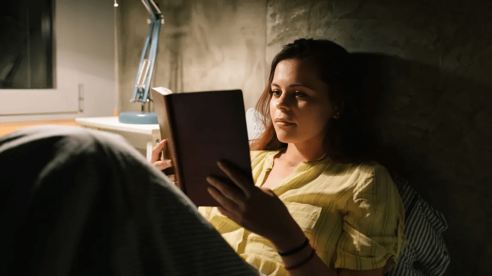 Woman in bed, reading a book by lamplight.