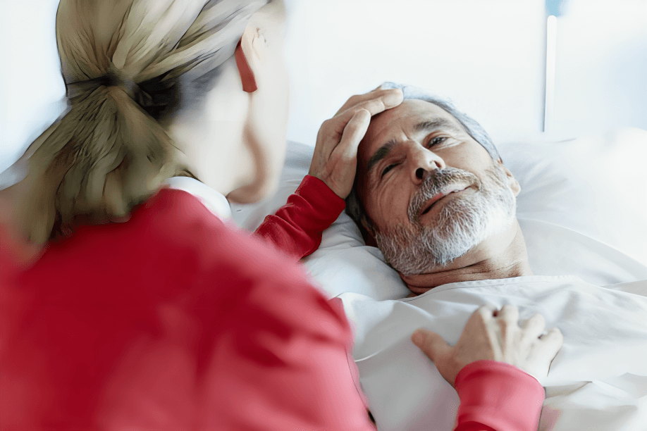 A woman comforts an older man lying in bed.