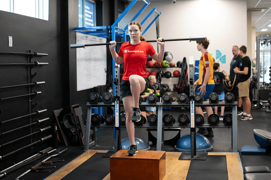 Girl doing box squats with a barbell in a gym.