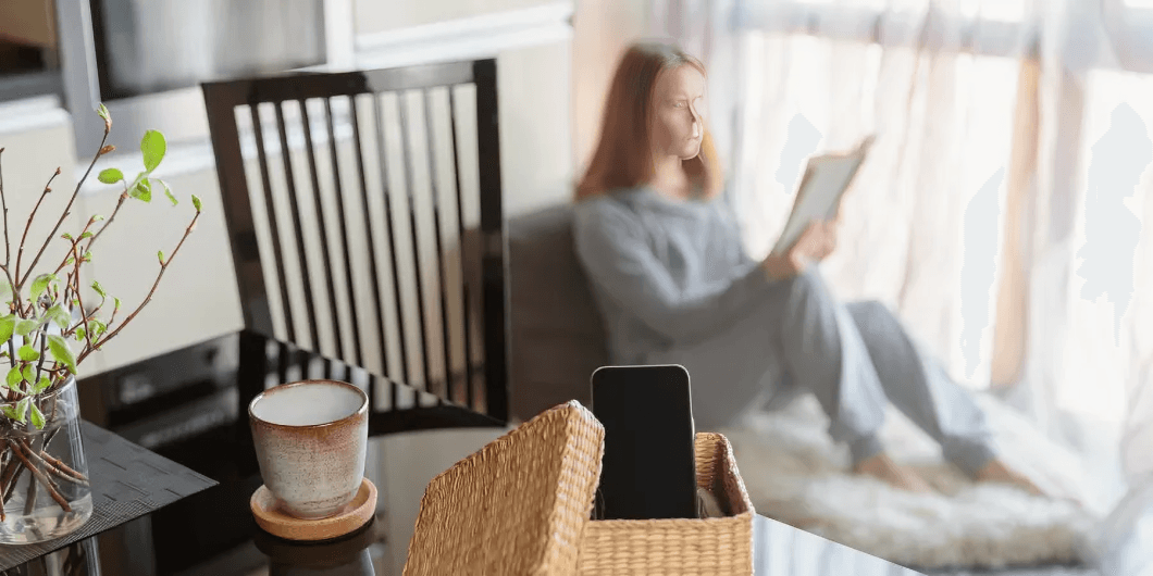 A smartphone in focus, blurred woman reading by window.