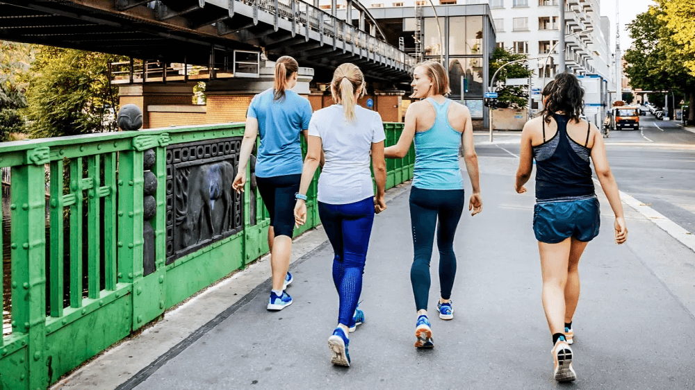 Four women walk together on a pedestrian bridge, backs to viewer.