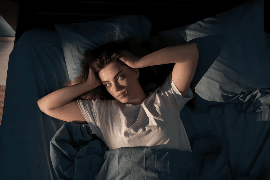 Woman awake in her bed, hands behind head, looking worried.