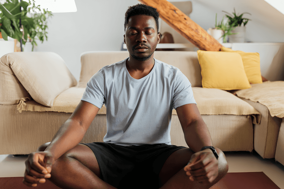 A man meditates in the lotus position on a mat at home.