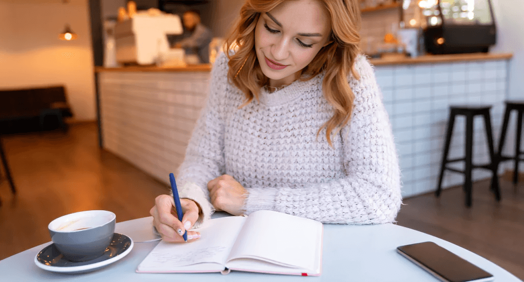 A woman with wavy hair writing in a notebook at a cafe.