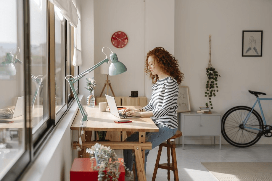 A young woman with curly red hair works on a laptop at a desk.