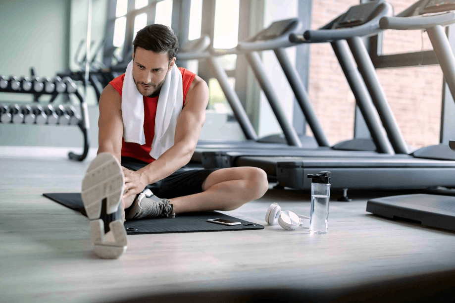 Man stretching in a gym with a towel and headphones.