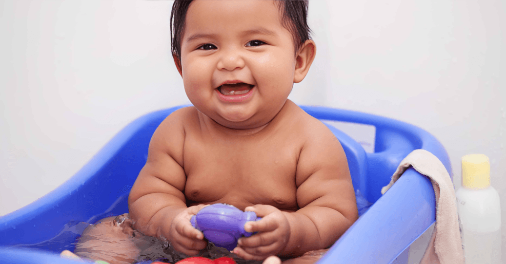 A cheerful baby with dark hair bathing in a blue tub.