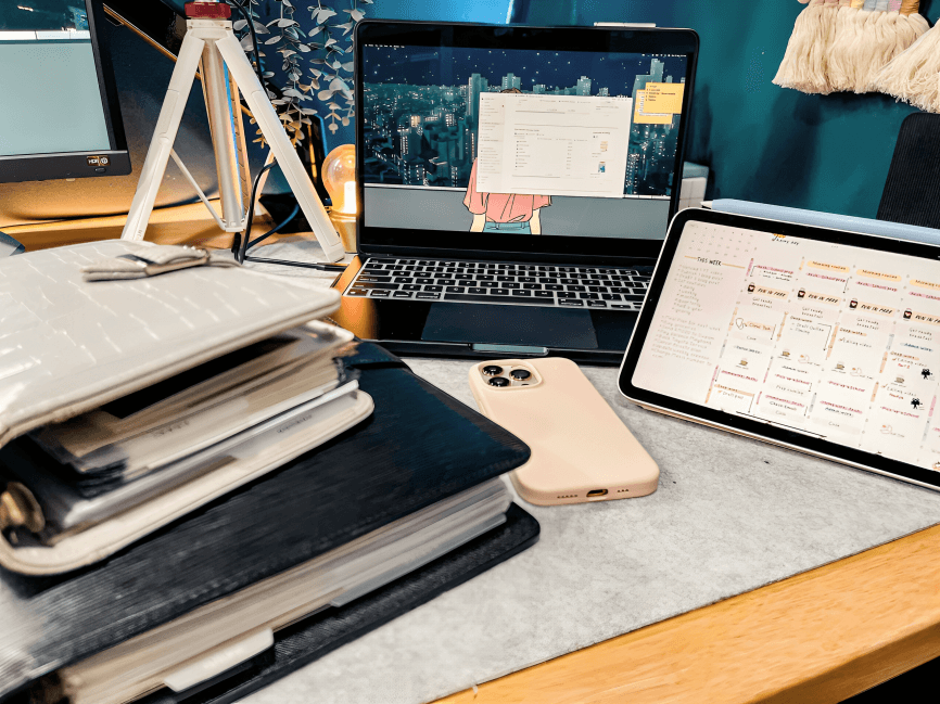 A laptop, tablet, and planner on a cluttered desk.