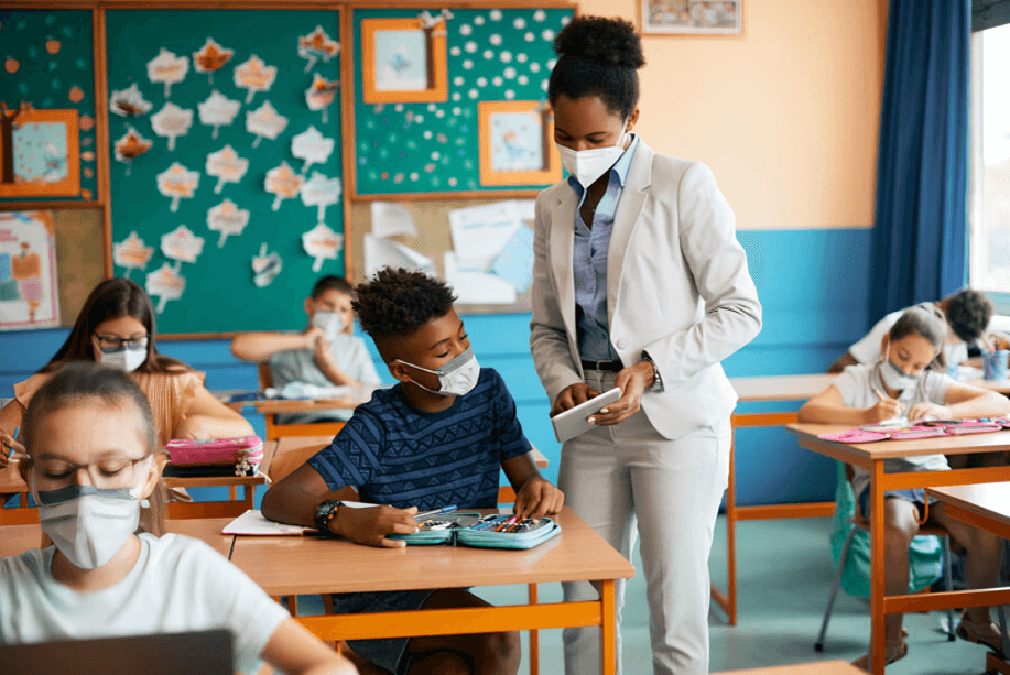 A masked teacher helps a masked student with a tablet in a classroom.