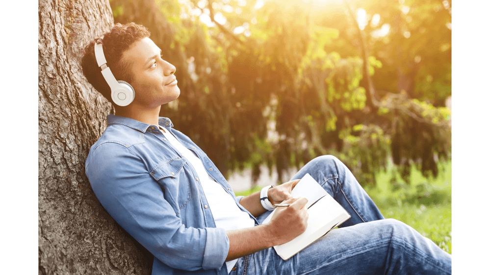 A man with headphones leaning on a tree, writing in a park.