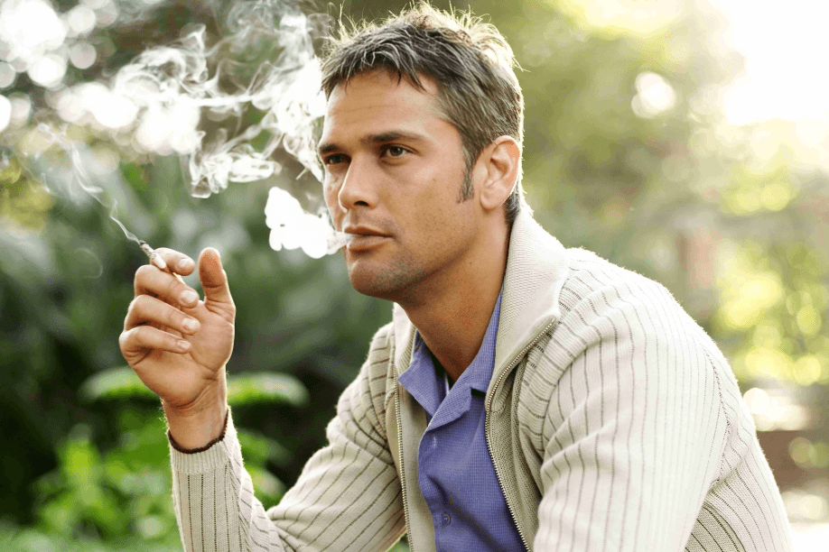 A man in a sweater smoking outdoors, exhaling smoke.