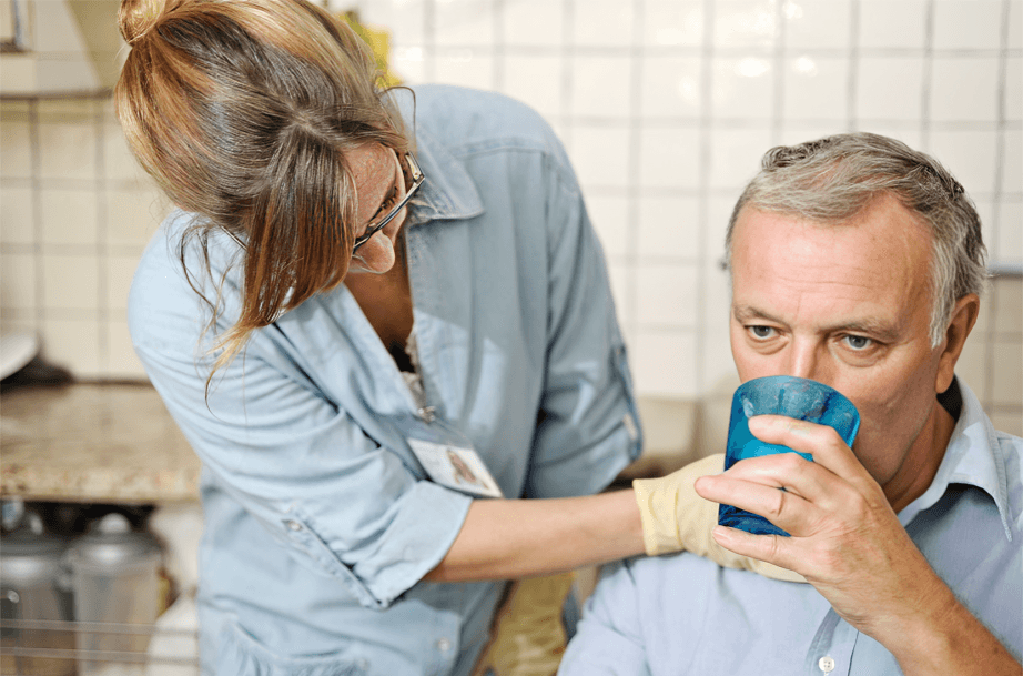 Caregiver assists elderly man drinking from a blue glass.