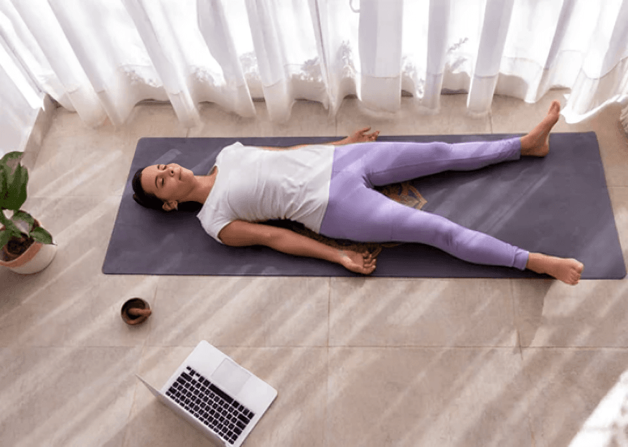 Woman lying on yoga mat near laptop and plant.