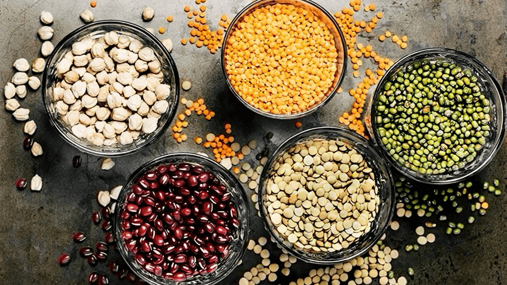 Five bowls of different legumes on a dark surface, top view.