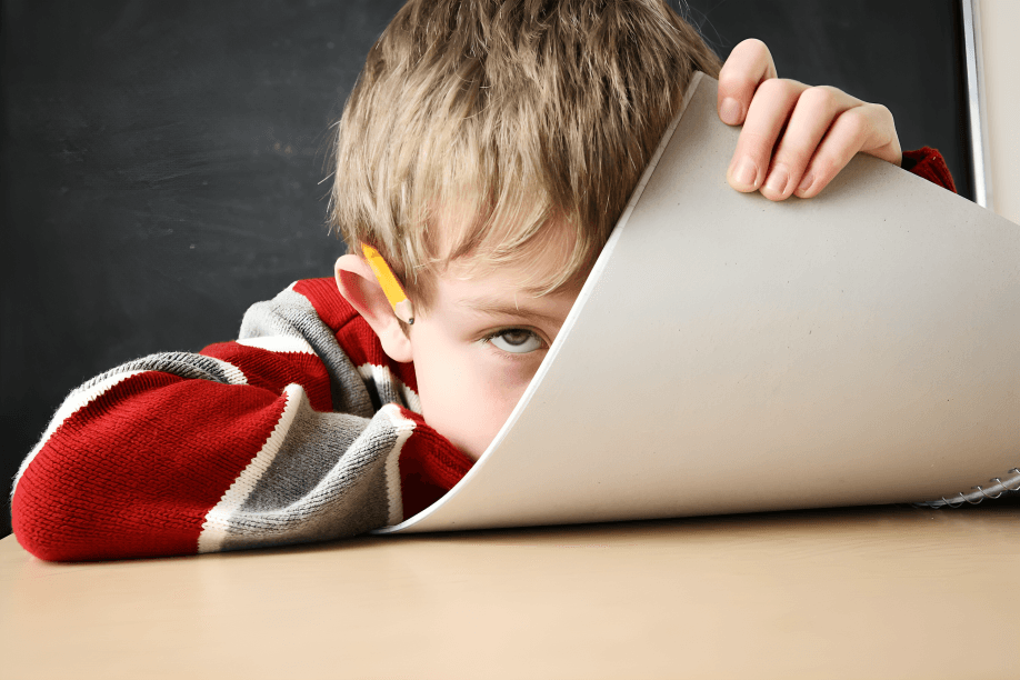 A boy peeks over a paper, holding a pencil.