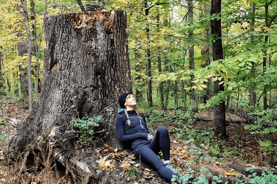 A woman sits peacefully by a large tree stump in the woods.
