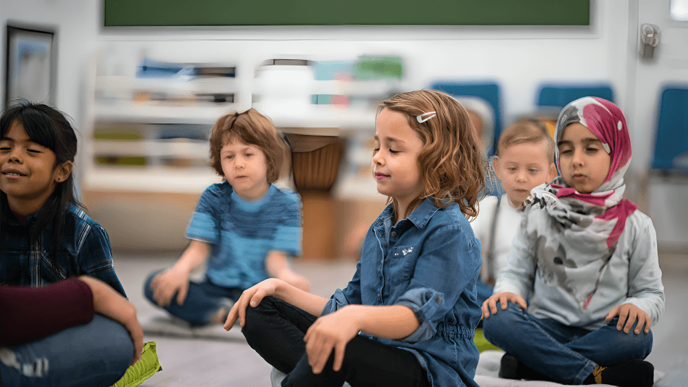 Five young children meditate on mats in a classroom.