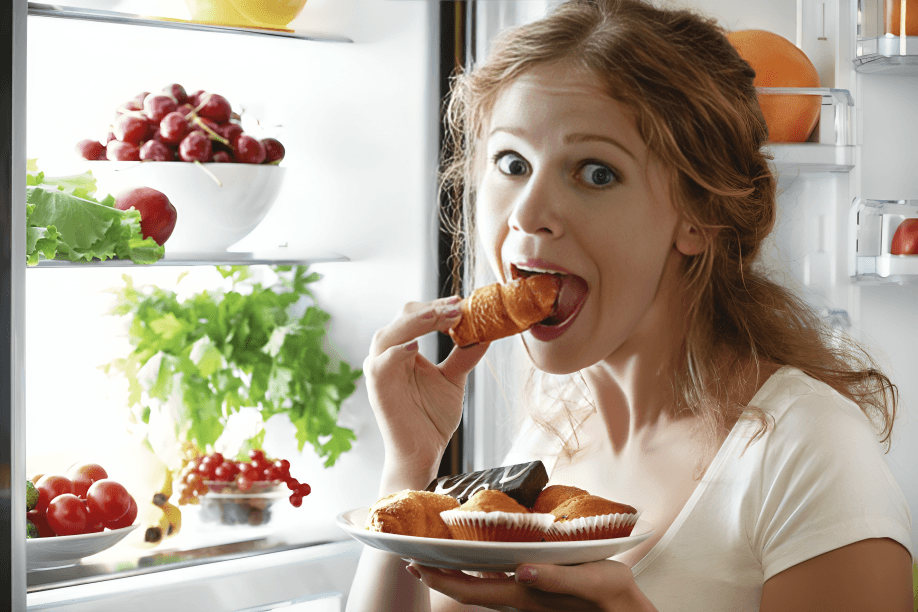 A woman eats pastries in front of an open fridge.