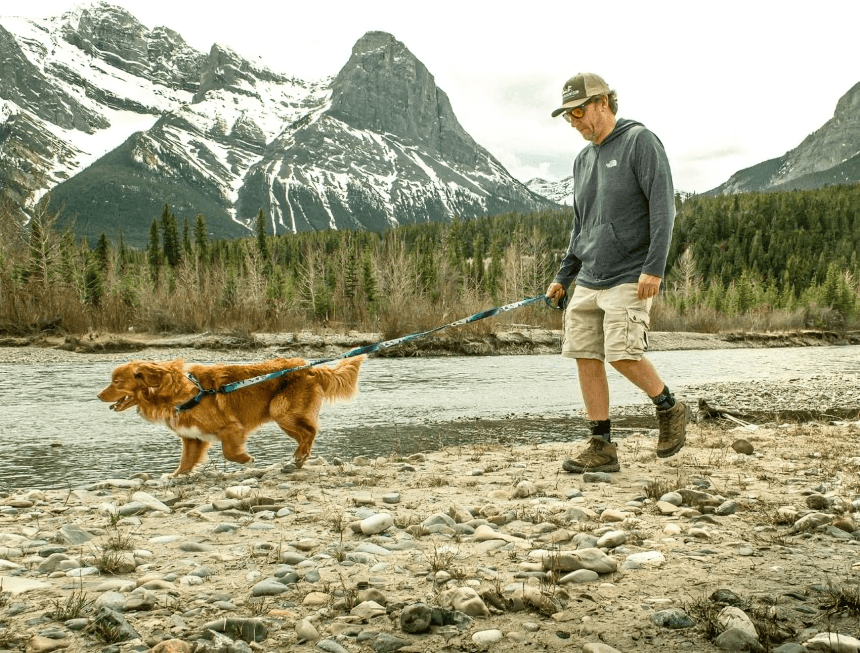 Man and dog walk by a river and mountains.